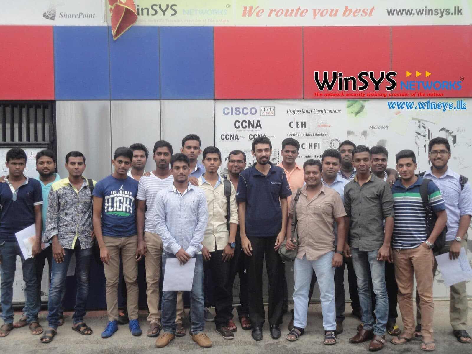A WinSYS student cohort outside the campus, under the CCNA and cyber security certification signage